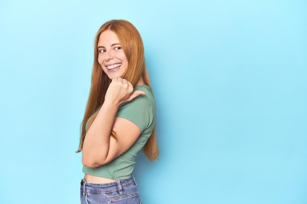 Redhead young woman on blue background points with thumb finger away laughing and carefree