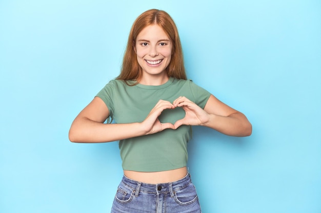 Photo redhead young woman on blue background smiling and showing a heart shape with hands