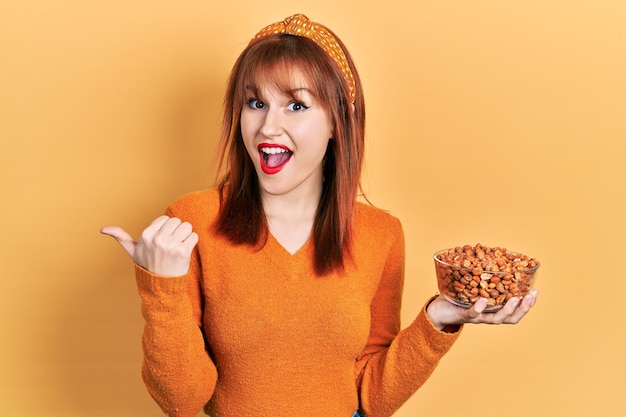 Redhead young woman holding peanuts pointing thumb up to the side smiling happy with open mouth