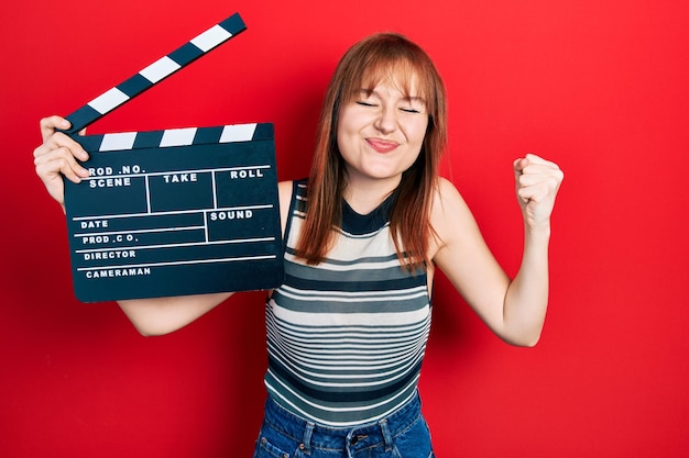 Redhead young woman holding video film clapboard screaming proud celebrating victory and success very excited with raised arm