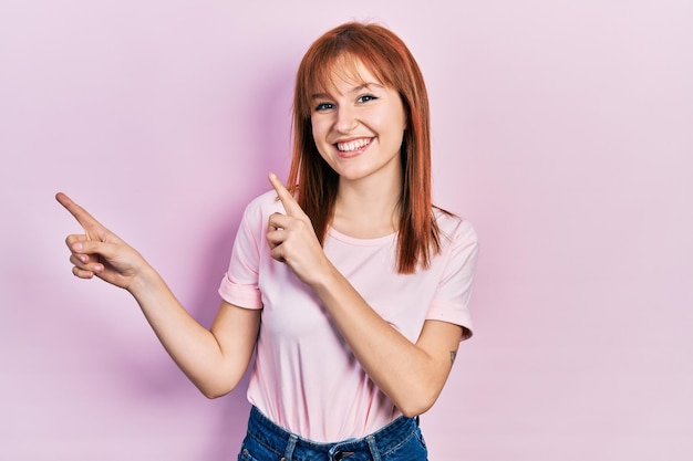 Redhead young woman wearing casual pink t shirt smiling and looking at the camera pointing with two hands and fingers to the side