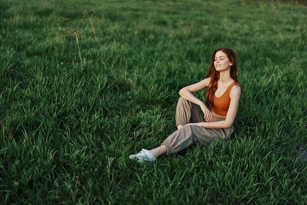 Photo the redheaded woman sits in the park on the green grass wearing an orange top green pants and sneakers and looks out at the setting summer sun high quality photo