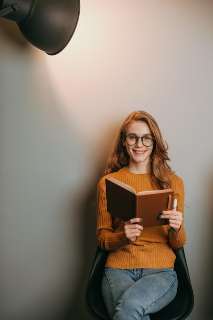 Redheaded woman sitting on a chair over white wall and reading a book posing in the spotlight busine