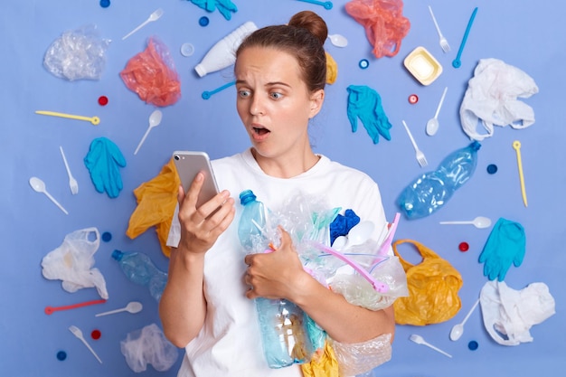 Reduce garbage pollution Cleaning up planet Shocked surprised woman wearing white shirt isolated on blue wall with garbage around looking at cell phone screen with open mouth