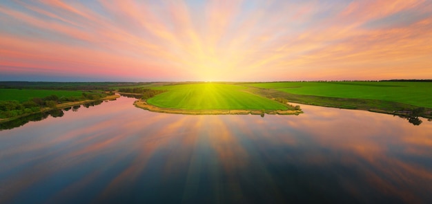 Reflection of a dramatic sunset sky in the calm water of a lake with a green shore. Panorama