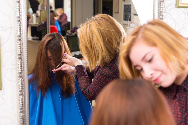 Reflection in Large Salon Mirror of Young Blond Stylist Cutting Hair of Brunette Client - Hair Stylist Trimming Bangs for Customer