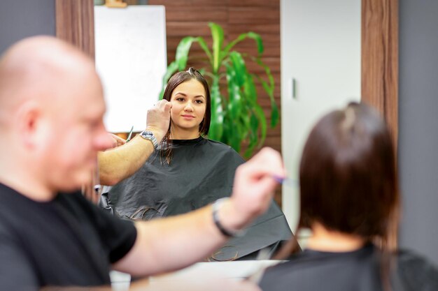 Reflection in the mirror of the young caucasian woman sitting and receiving a haircut by male