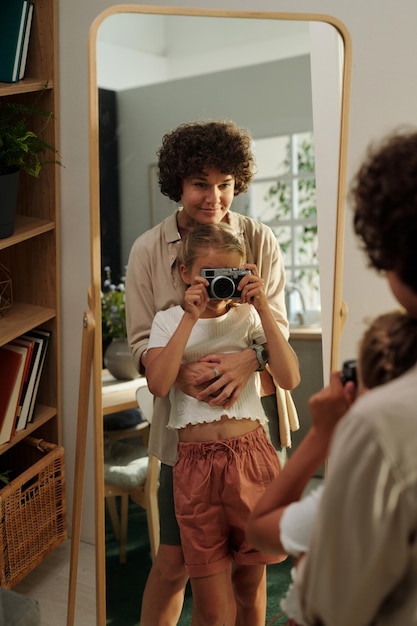 Reflection in mirror of young woman embracing her daughter with photocamera