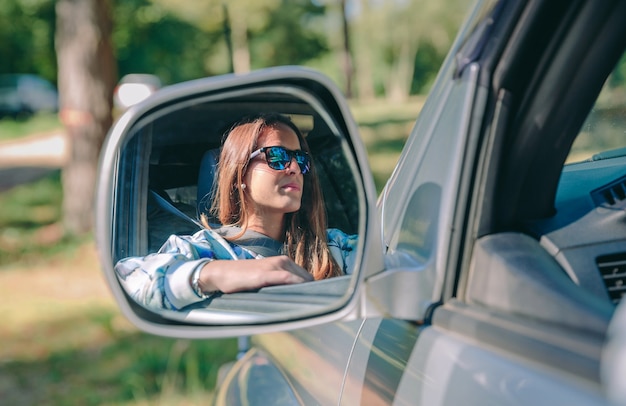 Reflection in side view mirror of young woman with sunglasses driving car over a nature background