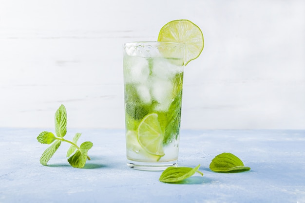 Refreshing cool drink with lime and mint on blue table. Summer lemonade or ice tea in a glass. Mojito cocktail with ice cubes.