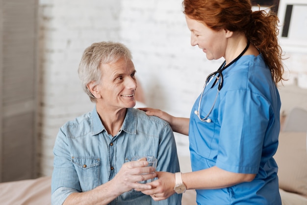 Refreshing drink. Charismatic motivated mindful woman making sure her patient feeling better while serving him a glass of water during the regular visiting at home