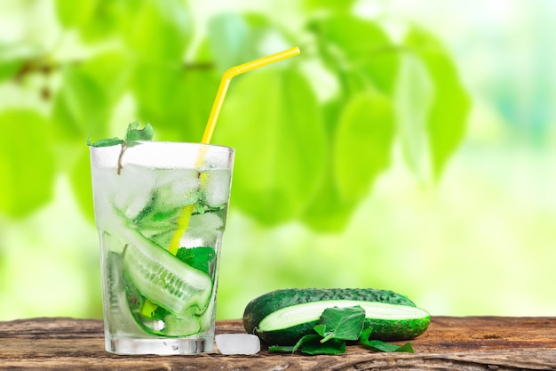 Refreshing drink with cucumber and mint in a glass with a straw next to ice cubes against a green background