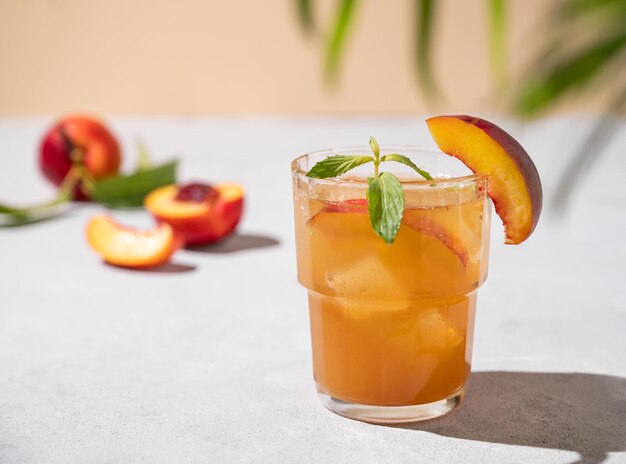 Refreshing peach tea with ice and mint Homemade cold healthy vegetarian drink on a light background with fresh fruits palm leaf and shadows close up