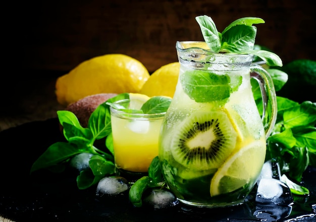 Refreshing summer cocktail with green basil lemon kiwi fruit and ice in a jug black background selective focus