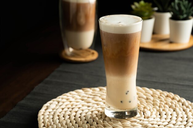 Refreshing summer drinks or beverages, consists of three layers, cold milk, coffee, and cold foam milk in a tall glass, on weaving placemat, grey placemat, dark brown wood table.