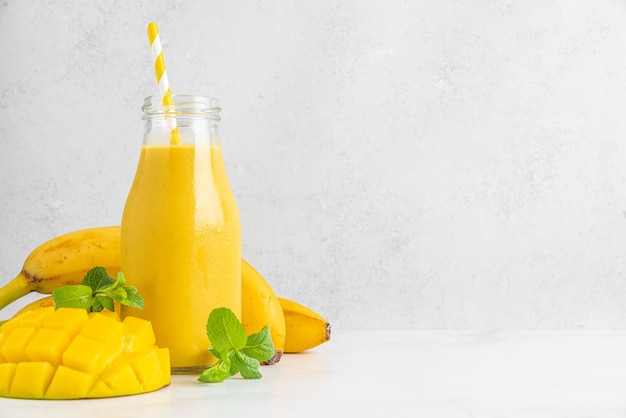 Refreshing tropical mango and banana smoothie in a glass bottle with fresh fruit mint and straw on white background