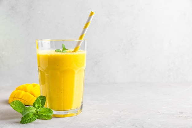 Refreshing tropical mango smoothie in a glass with fresh fruit mint and straw on gray concrete background