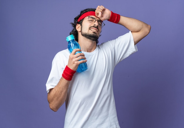 Regretted with closed eyes young sporty man wearing headband with wristband holding water bottle putting hand on forehead 