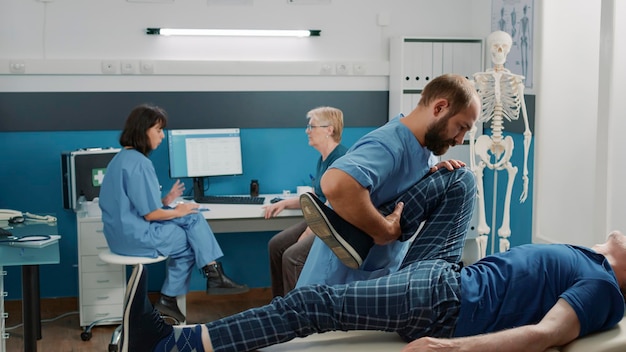 Rehab specialist using legs raise exercise to stretch muscles, giving assistance to old patient at recovery appointment. Osteopath doing rehabilitative physiotherapy with elder man.