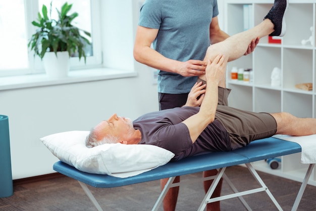 Rehabilitation activity. Good looking aged man raising his leg while doing rehabilitation activity