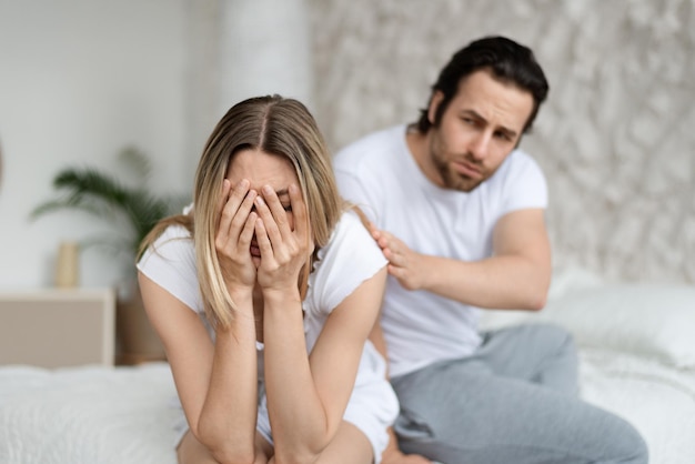 Relationship of family problems in couple quarrel concept Loving caucasian man calming his crying wife sitting on bed