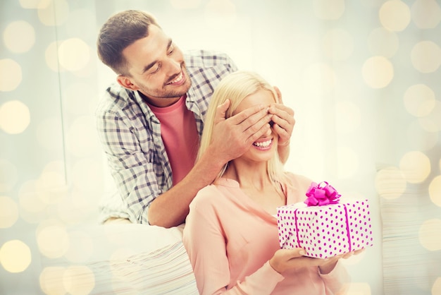 relationships, love, people, birthday and valentines day concept - happy man covering woman eyes and giving gift box at home over holidays lights background