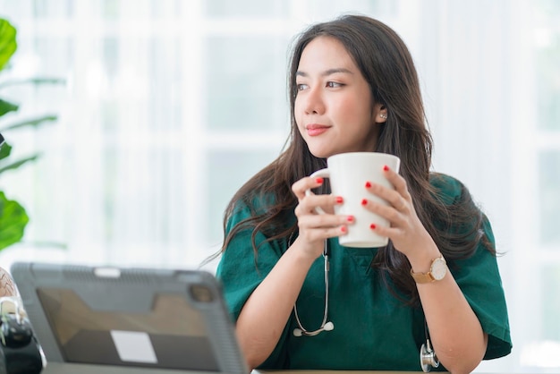 Relax after work asian female doctor hand hold hot coffee drink cup casual relax after working hard telemedicine in clinic while wear uniform