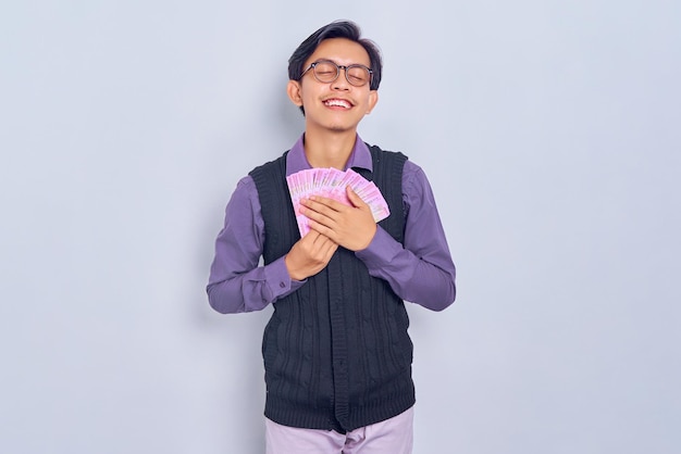 Relax and smiling young Asian man in purple shirt outfit holding salary cash money in rupiah banknotes isolated on white background People lifestyle concept