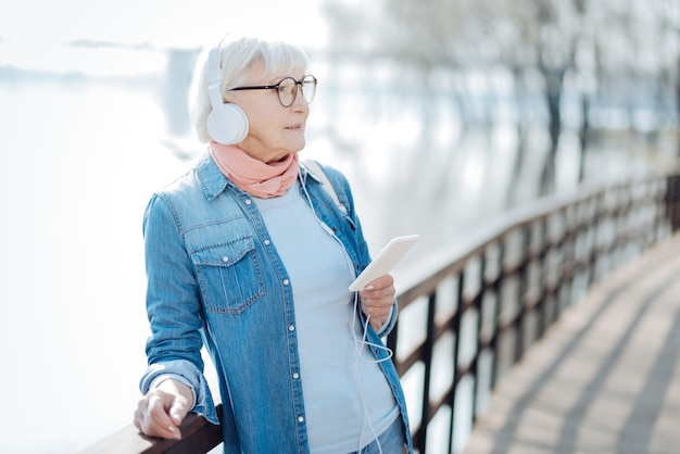 Relaxation. Exuberant aged woman listening to music while walking in the park