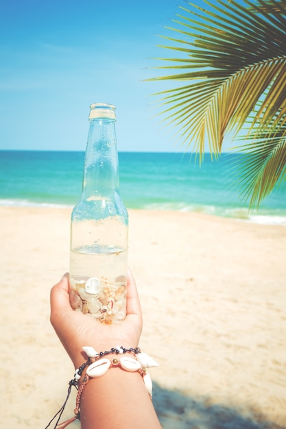 Relaxation and Leisure in summer - Young tanned woman hand holding bottle with seashell at tropical beach in summer. vintage color tone effect