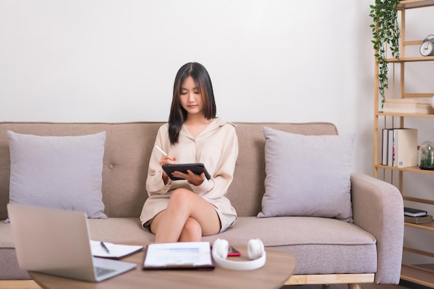 Relaxation lifestyle concept Asian woman sitting on sofa and writing on tablet to working at home