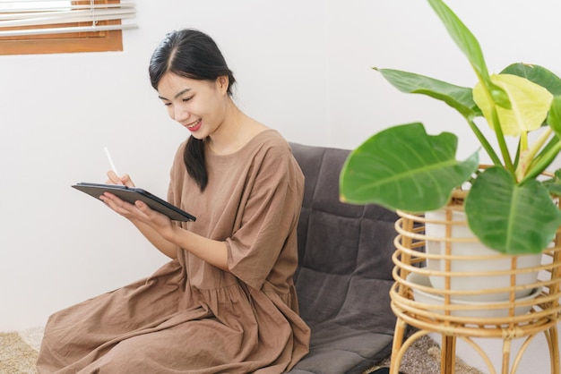 Relaxation lifestyle concept Young Asian woman writing on tablet while sitting in living room