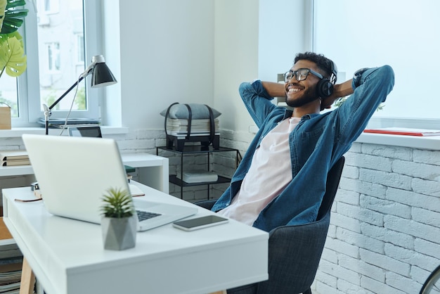 Relaxed African man in headphones holding hands behind head while sitting at working place