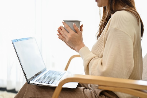 Relaxed Asian female sits on armchair in the living room holding a coffee mug and using laptop