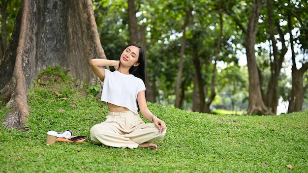 Relaxed Asian female sitting under the tree stretching and massaging her neck