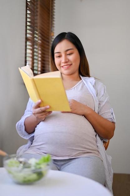 Relaxed Asian pregnant woman reading a book at a dining table preparing to be a mother