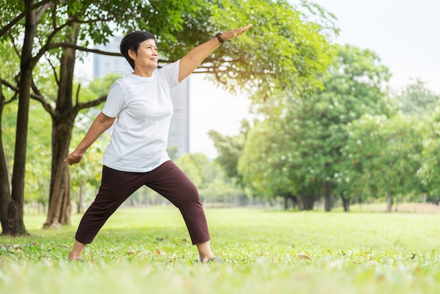 Relaxed Asian Senior woman in white cloth doing stretching workout her arms at park. Smiling Elderly Thai Female enjoying exercising at nature outside