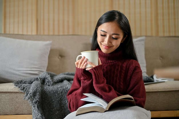 A relaxed Asian woman enjoys her morning coffee while reading a book in her living room