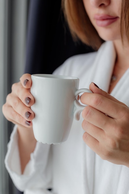 Relaxed Asian woman wearing bath robe drinking coffee in hotel room in morning