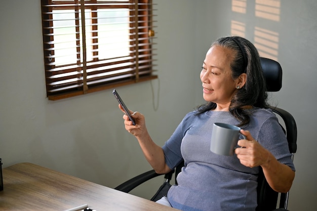 Relaxed Asianaged businesswoman sipping coffee and using her phone at her desk