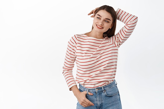 Relaxed beautiful girl hold hands behind head, standing in casual resting pose, looking aside at copyspace promotional text and smiling, standing over white background