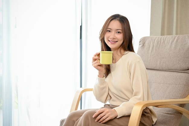 Relaxed and beautiful young Asian woman is on her armchair while having a coffee