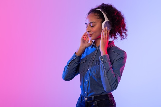 Relaxed black lady enjoying music in headphones denim attire colorful backdrop