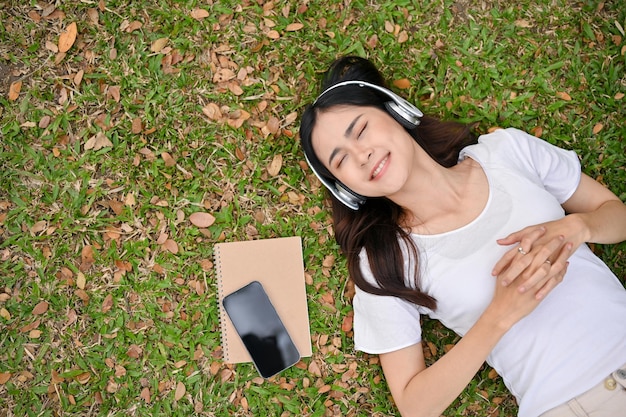 Relaxed and chilling young Asian woman listening to music smiling eyes closed and lying on grass