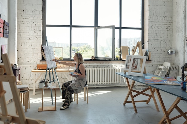 Relaxed female artist in apron holding palette and drinking wine while thinking of painting