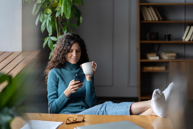 Relaxed female employee drinking coffee and using cellphone at workplace with legs on office table