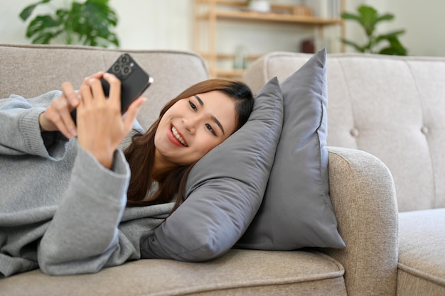 Relaxed and happy Asian female using smartphone while laying on her comfortable sofa