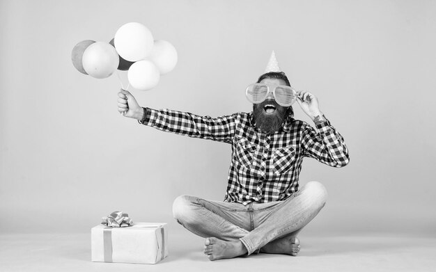 Relaxed happy birthday guy looks cheerful bearded man feel the joy man with gift box celebrating birthday mature hipster with bright colorful balloons Crazy funny guy