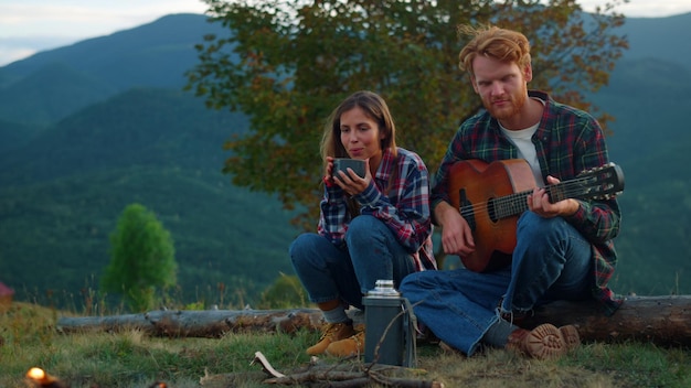 Relaxed lovers play guitar on nature view Hikers couple spend vacation outdoors close up Young family enjoy camping on green hill mountains Happy tourists play guitar by campfire Leisure concept