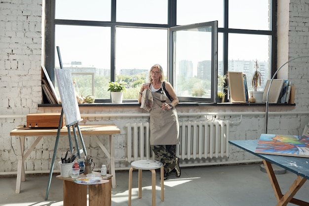 Relaxed mature female artist in apron standing at window and drinking wine in art studio